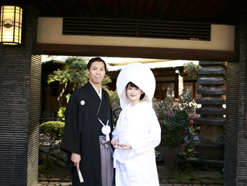 一松の黒塀の前に立つ和装姿の花嫁と花婿